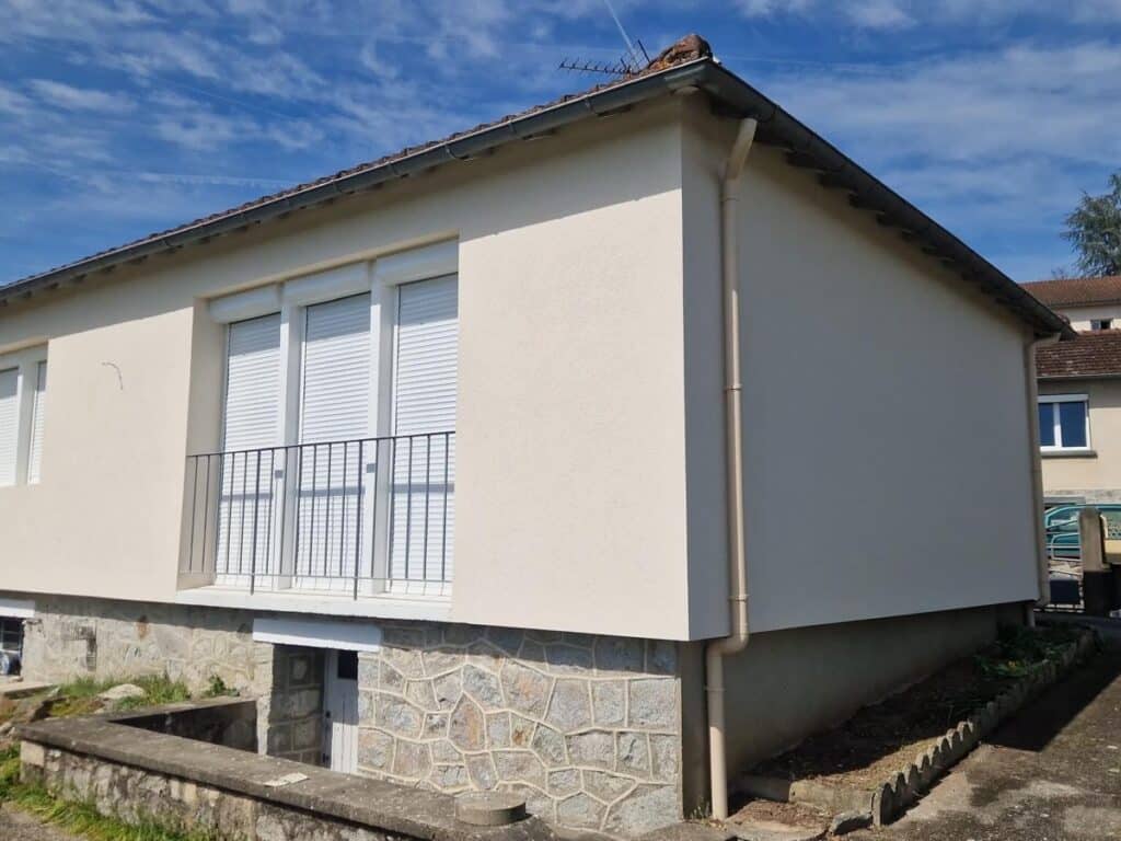 Façade de maison beige avec soubassement en pierre Maison à façade beige et soubassement en pierre avec volets roulants blancs et balcon sous un ciel bleu.