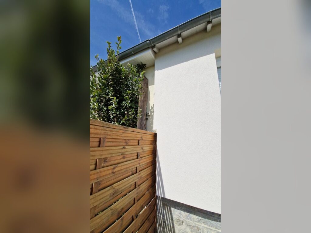 Façade Maison : Mur Blanc avec Clôture en Bois Moderne Mur de maison crépi blanc, clôture en bois horizontale et arbuste verdoyant sous un ciel bleu ensoleillé.